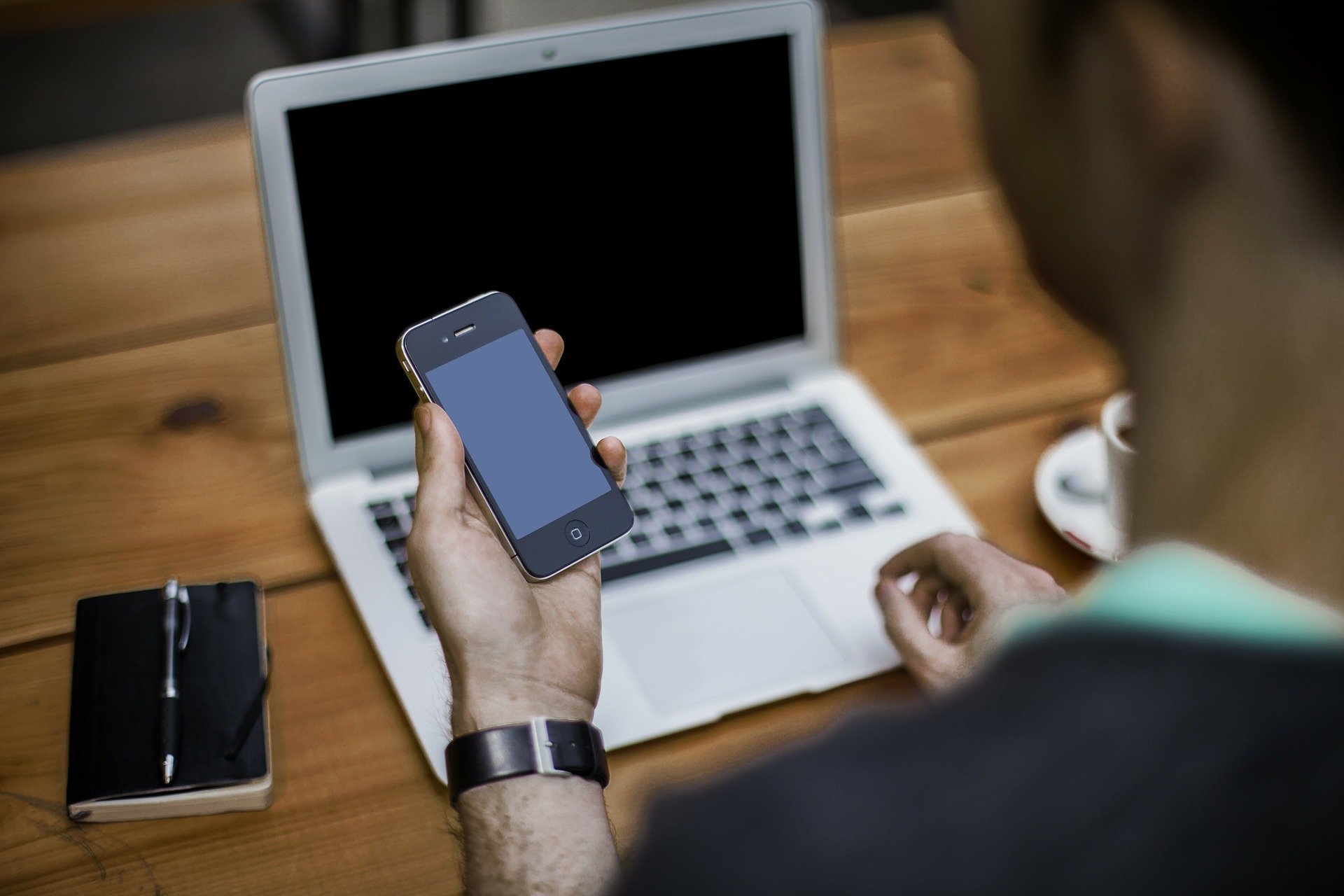 A man sitting in front of a laptop and an mobile phone in his hand at a deak.