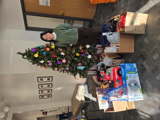Image of an employee in front of a christmas tree with all the gifts that were collect for Community Threads holiday program