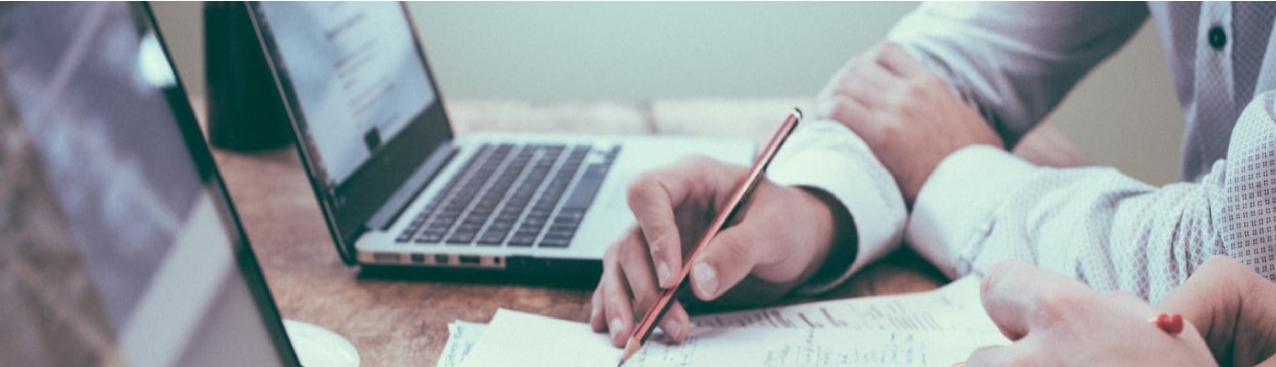 An image of a couple laptops and a persons hand holding a pencil writing on paper